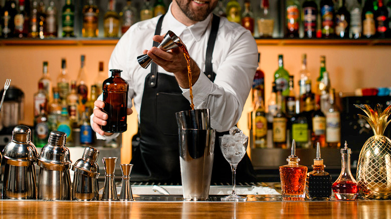 various steel shakers and bottles stand on the bar counter, and man bartender gently pours an alcoholic drink from jigger in the shaker cup