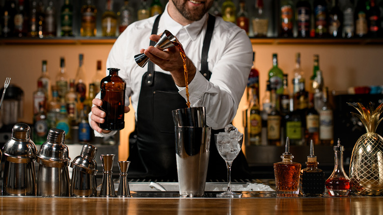 various steel shakers and bottles stand on the bar counter, and man bartender gently pours an alcoholic drink from jigger in the shaker cup